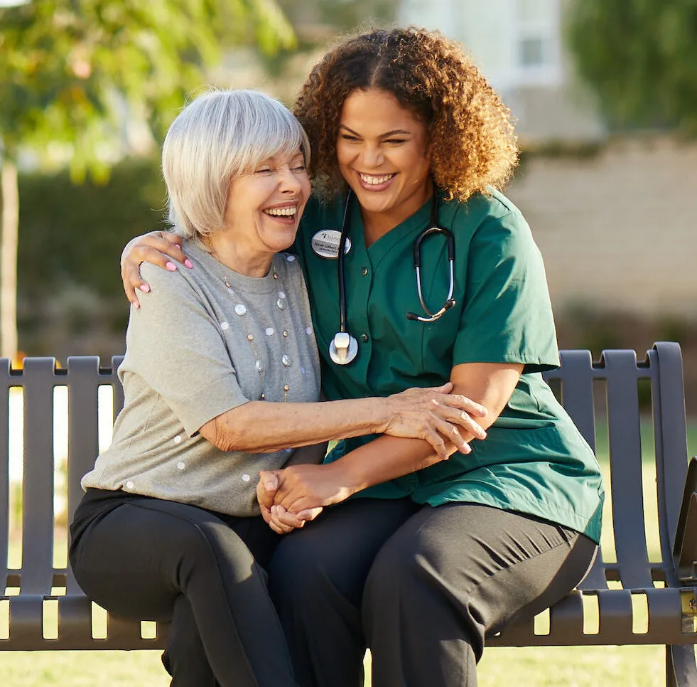 Caregiver is laughing with the senior lady on a bench