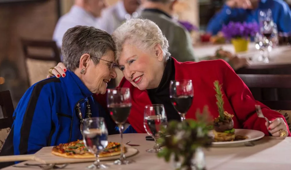 Two elegant dressed senior ladies embracing each others while having food in the dining room