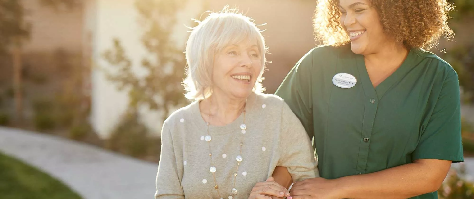 Caregiver and senior lady walking along path, smiling at each other