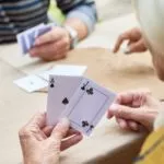 An elderly group of people playing cards, with one person holding an Ace of clubs and a Jack of spades.